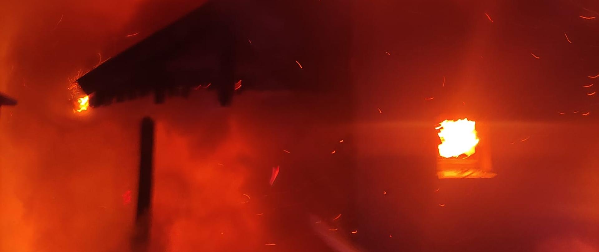 Zdjęcie nocne pokazuje pożar dachu budynku. Widoczne są płomienie na połaci dachowej oraz strumień wody podawany przez strażaków w celu ugaszenia ognia.