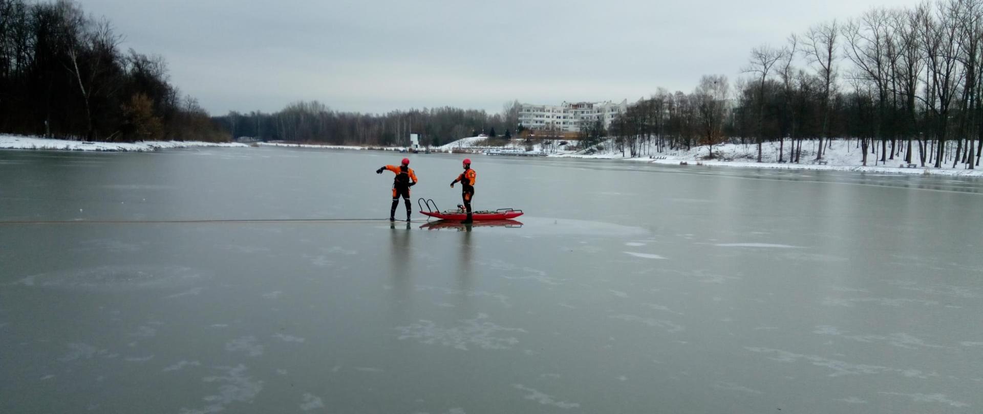 Zdjęcie przedstawia zalew kielecki na którego pokrywie lodowej stoi dwóch ratowników zabezpieczonych w specjalne kombinezony oraz sanie lodowe. 