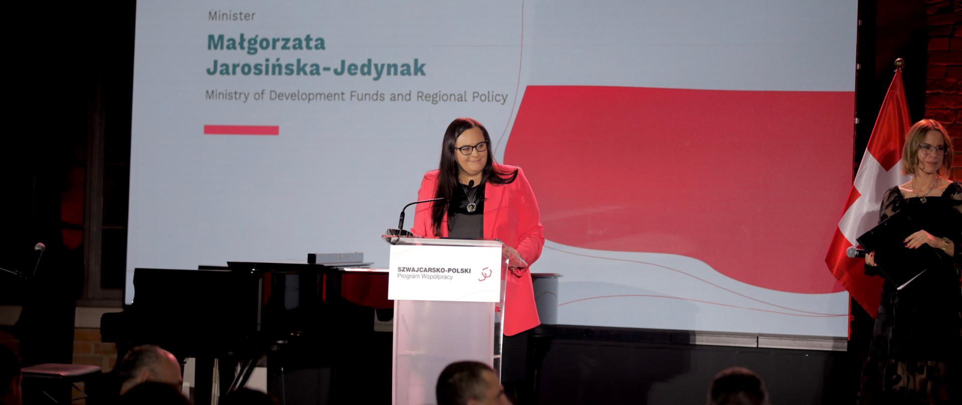 Minister Małgorzata Jarosińska-Jedynak w mównicy na scenie. Za nią flagi Polski i Szwajcarii. Obok prowadząca konferencję.