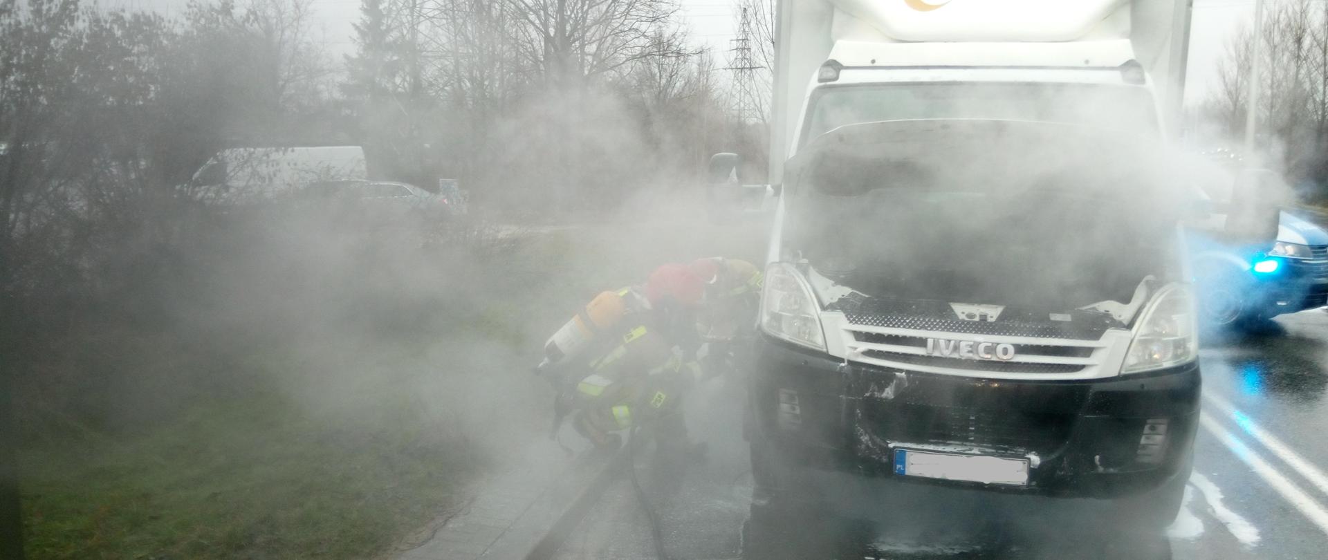 Zdjęcie przedstawia samochód dostawczy: stoi na pasie jezdni i ma podniesioną klapę nad komorą silnika z której wydobywa się dym i para wodna. Przed autem rozciągnięty na jezdni leży wąż szybkiego natarcia, a dwaj strażacy z lewej dogaszają pożar.