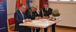 Przy wspólnym stole siedzi dwóch strażaków OSP w Brzezinach oraz przedstawiciel Zarządu WFOŚiGW w Rzeszowie. Wszyscy podpisują dokumenty. W tle ściana oraz banery starostwa powiatowego w Ropczycach.