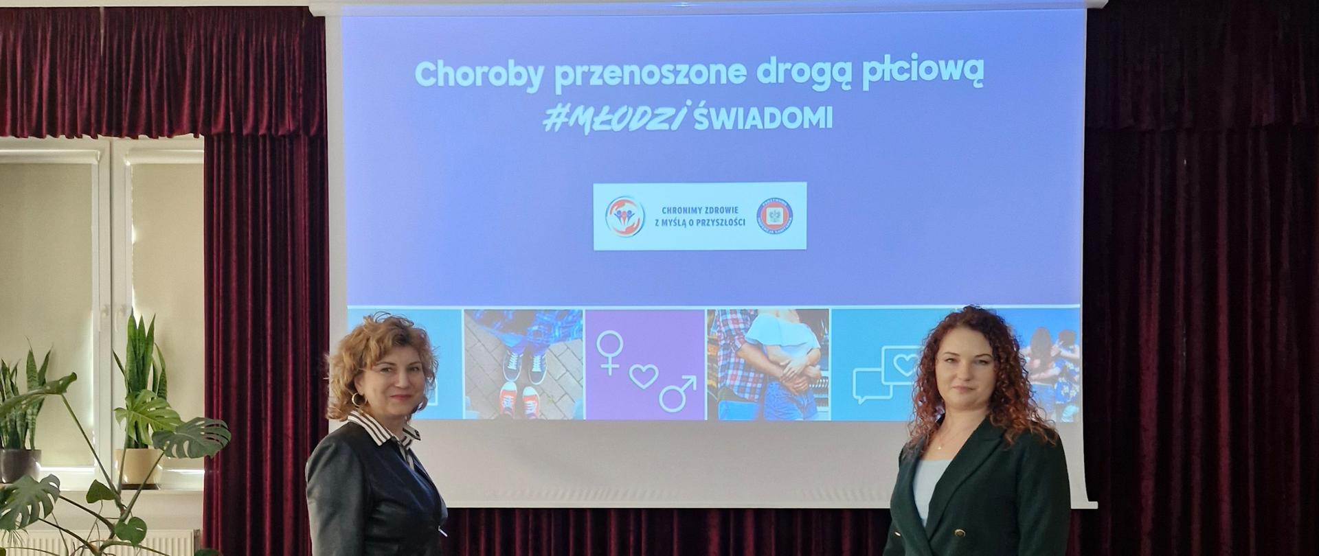 Dwie przedstawicielki Państwowego Powiatowego Inspektora Sanitarnego w Garwolinie na tle slajdu z treścią "Choroby przenoszone drogą płciową "MłodziŚwiadomi"