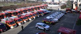Na placu w trzech rzędach stoi kilkadziesiąt samochodów strażackich: osobowych, dostawczych oraz ciężarowych. W tle widok miasta: park oraz zabudowania, w tym bloki