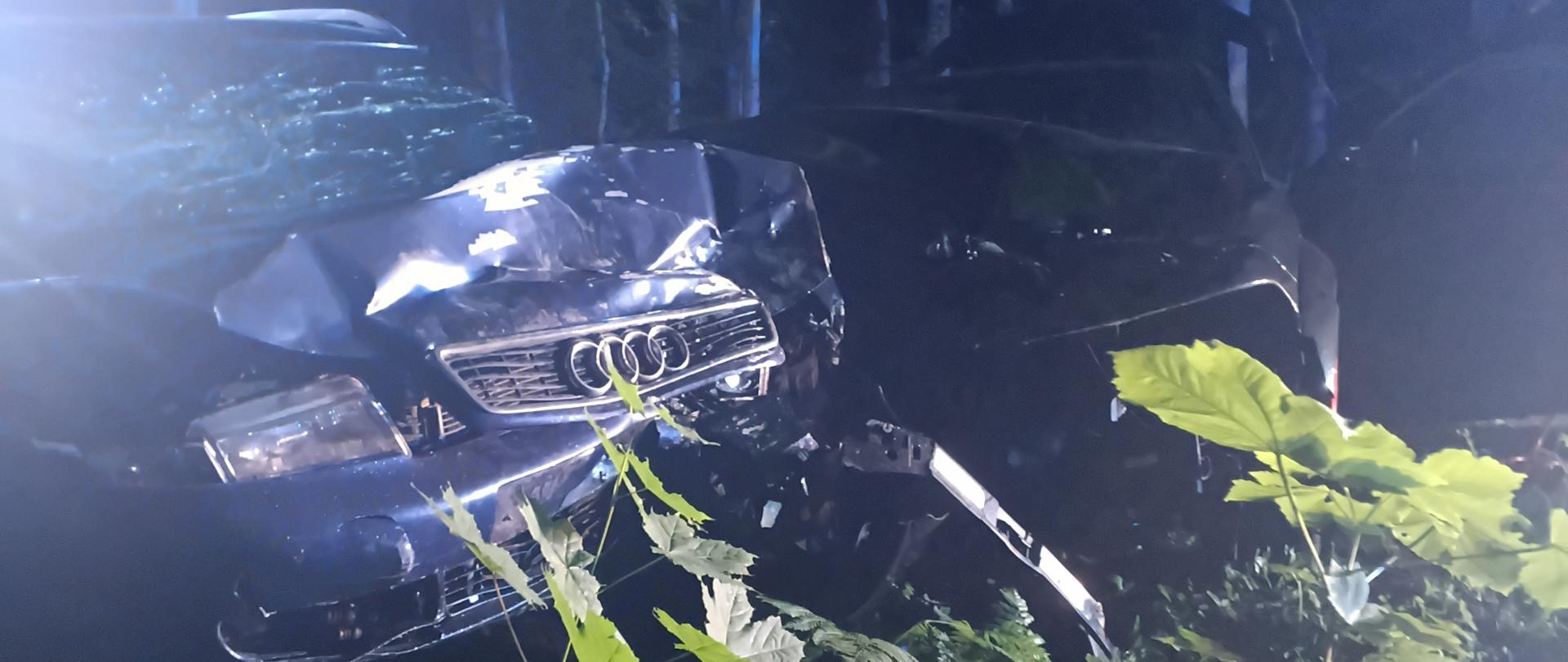Dwa pojazdy osobowe z mocno uszkodzonymi przodami znajdują się poza jezdnią, w przydrożnych zaroślach. Zdjęcie wykonano nocą podczas działań służb ratowniczych.