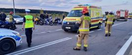 Zdjęcie przedstawia zdarzenia drogowe z udziałem motocykli. Na pierwszym Planie stoi strażak w ubraniu bojowym, dalej widać karetkę oraz inne służby. W tle widać ustawione sportowe motocykle.