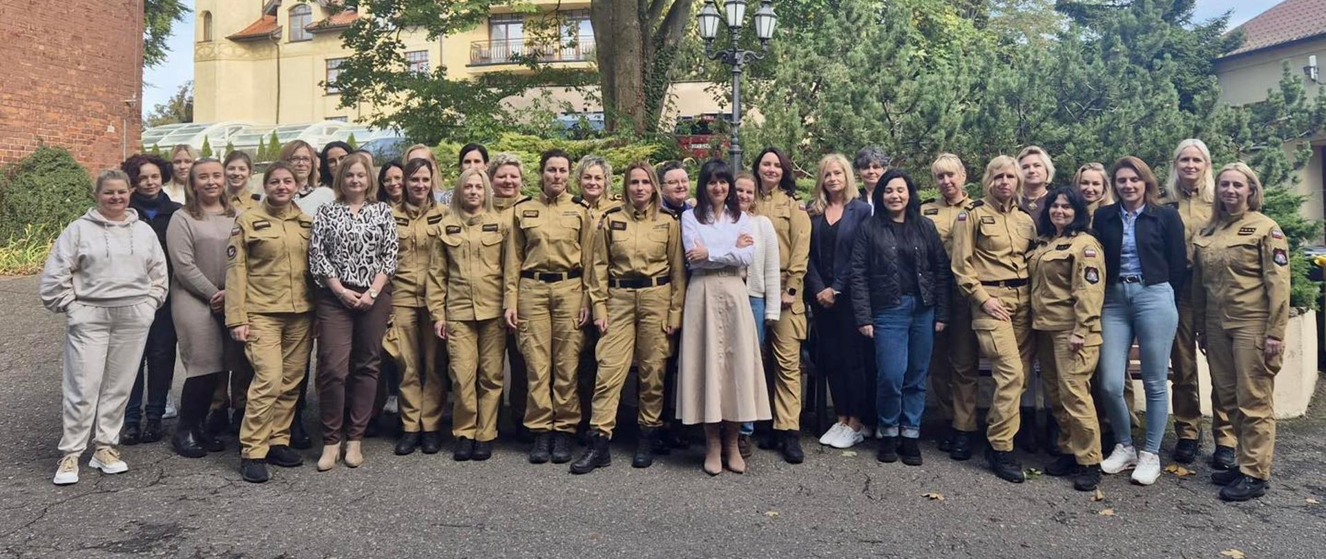 Na zdjęciu widać grupę około trzydziestu kobiet stojących na zewnątrz, na tle drzew i budynków. Część z nich ma na sobie piaskowe mundury, a część ubrana jest w codzienne ubrania – spodnie, kurtki, swetry i bluzy. Kobiety stoją w dwóch rzędach i patrzą w stronę aparatu, większość z nich się uśmiecha. Tło stanowi zielone otoczenie – drzewa z zielono-żółtymi liśćmi, krzewy oraz fragmenty zabudowań z jasnymi ścianami i czerwonymi dachami. Pogoda jest słoneczna, niebo jest jasne z kilkoma chmurami.