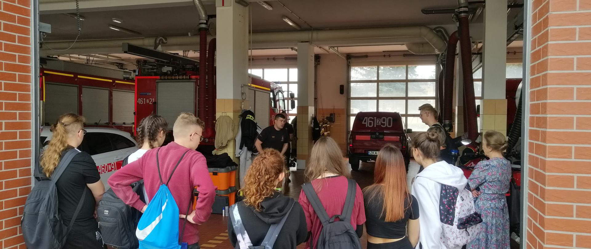 Zdjęcie przedstawia uczniów na terenie garażu JRG podczas szkolenia. w tle widać samochody PSP oraz strażaka który prowadzi wykłady.