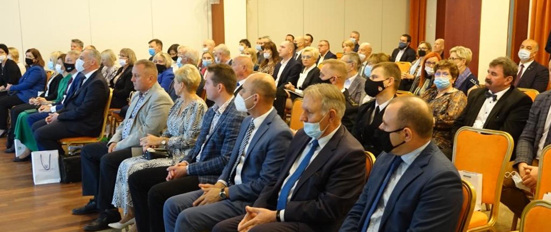 na zdjęciu około 50 osób siedzących na sali na krzesłach, mężczyźni ubrani w marynarki, kobiety w kostiumy