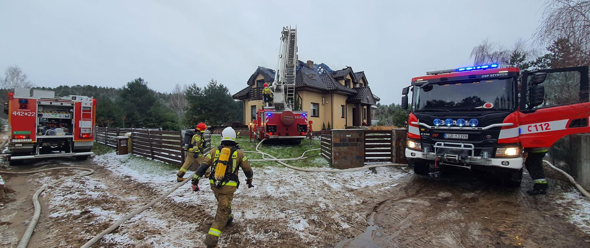 Zdjęcie przedstawia dom wolnostojący w kolorze beżowym z ciemną dachówką, przy którym pracują strażacy wykorzystując 3 czerwone wozy bojowe.