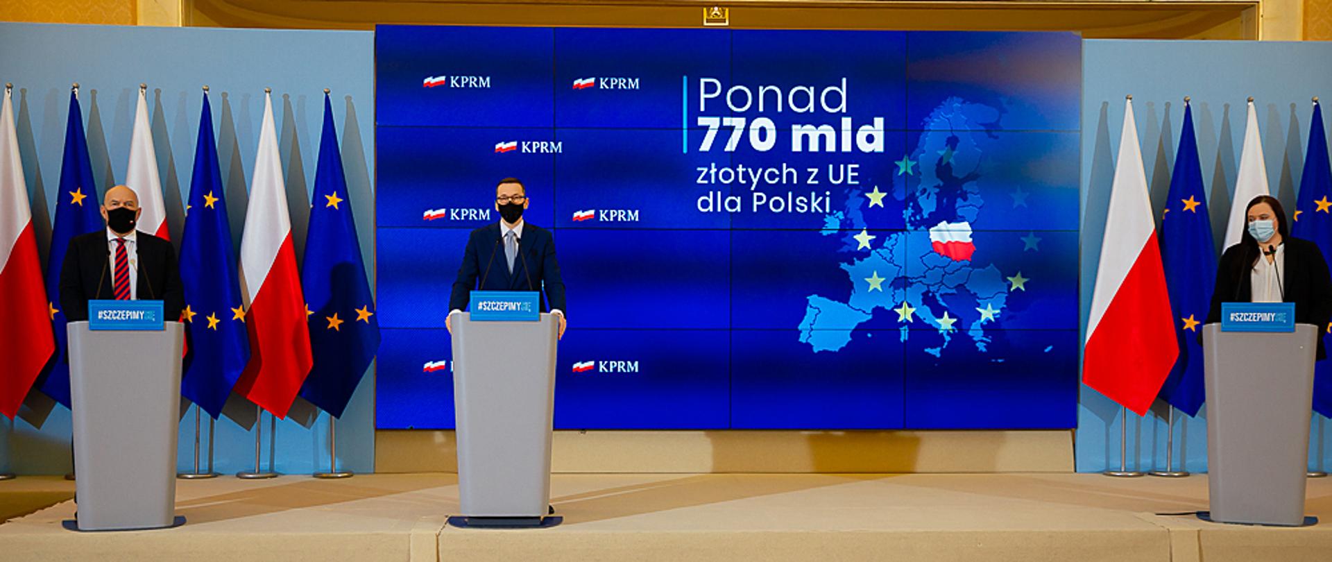 W mównicach trzy osoby. Od lewej minister Tadeusz Kościński, premier Mateusz Morawiecki oraz wiceminister Małgorzata Jarosińska-Jedynak. Za nimi flagi Polski i UE oraz napis "Ponad 770 mld złotych UE dla Polski".