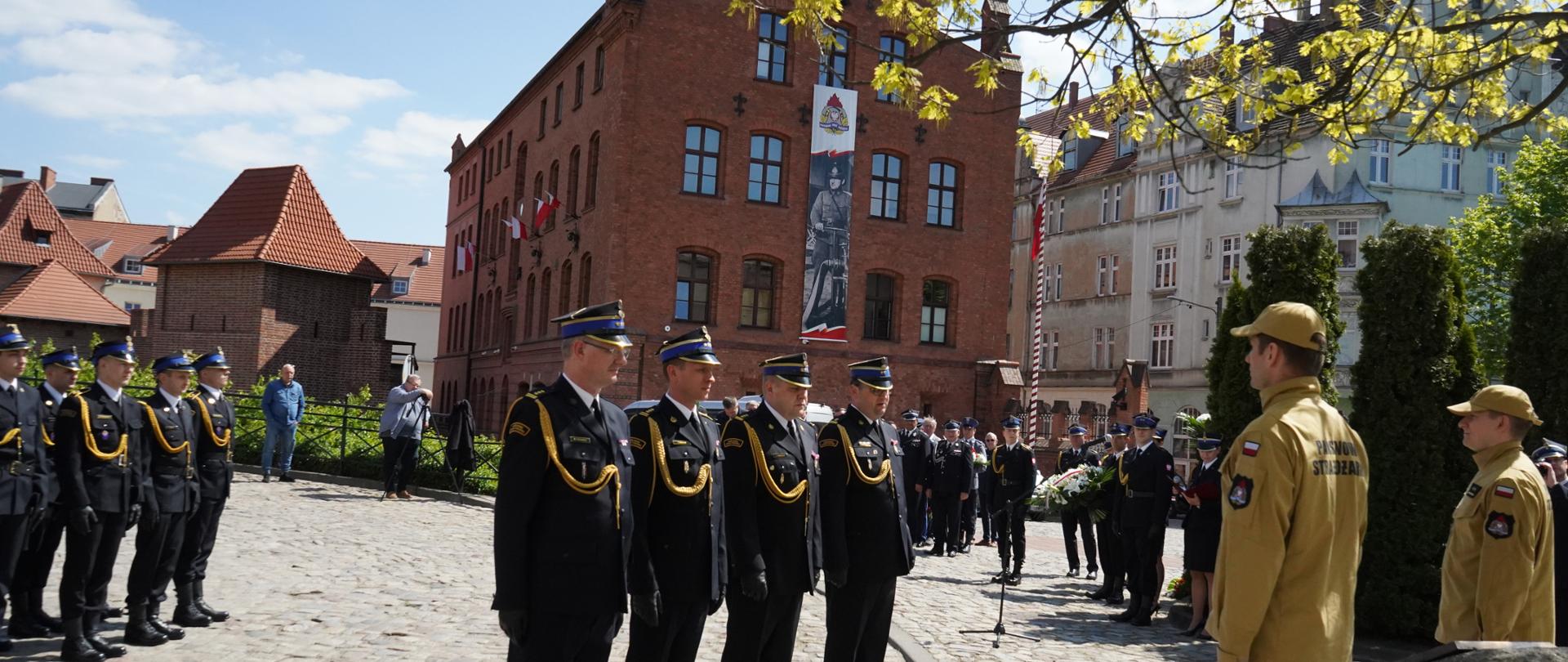 czterech strażaków stoi na baczność przed kamieniem, pomnikiem, w tle grupa strażaków i budynek strażnicy