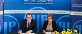 2 osoby na tle banneru w kolorze niebieskim z logo i napisem Państwowa Inspekcja Pracy. Osoby siedzą przy stole podpisują porozumienie. Od lewej strony: Pan dr Karol Łapiński p.o. Okręgowego Inspektora Pracy w Białymstoku, Pani dr n. med. Maria Siewko – Podlaski Państwowy Wojewódzki Inspektor Sanitarny w Białymstoku.