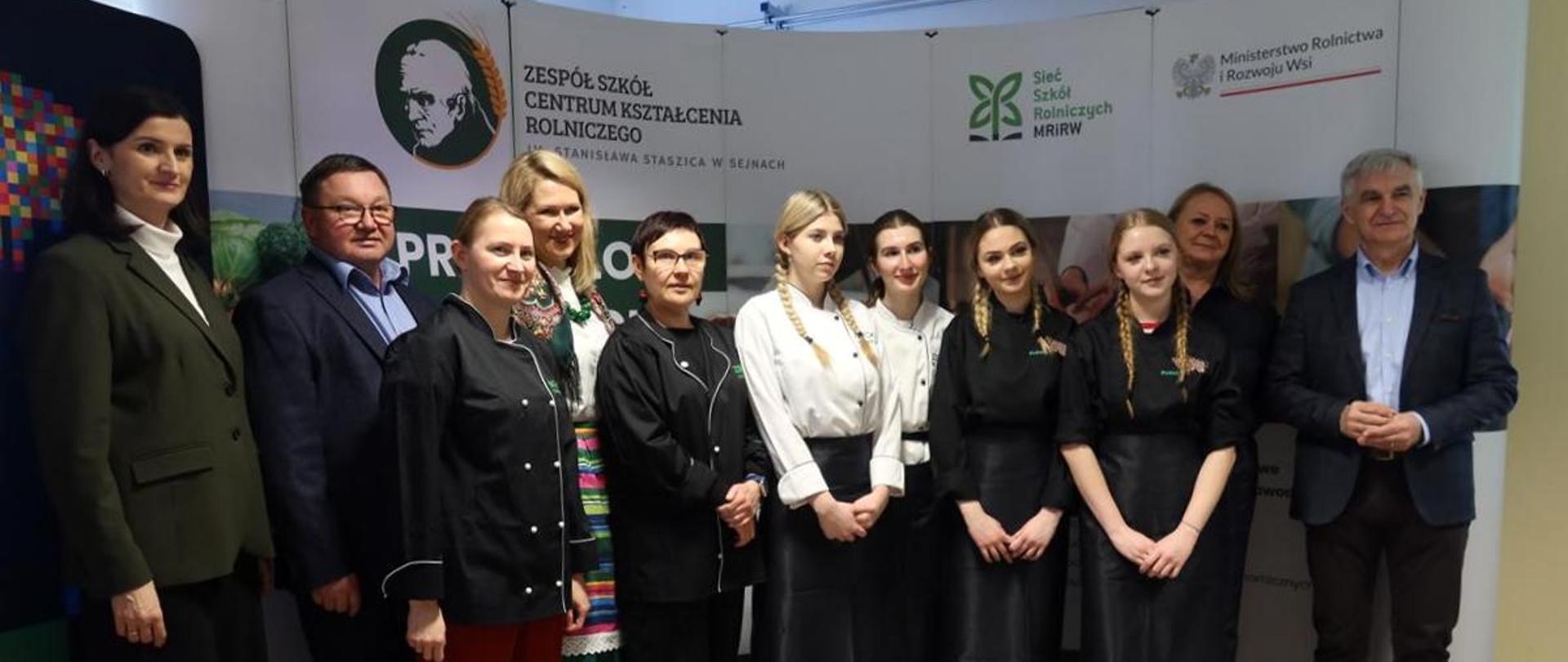 Cztery uczennice w strojach kucharzy oraz grupa dorosłych pozują do zdjęcia.