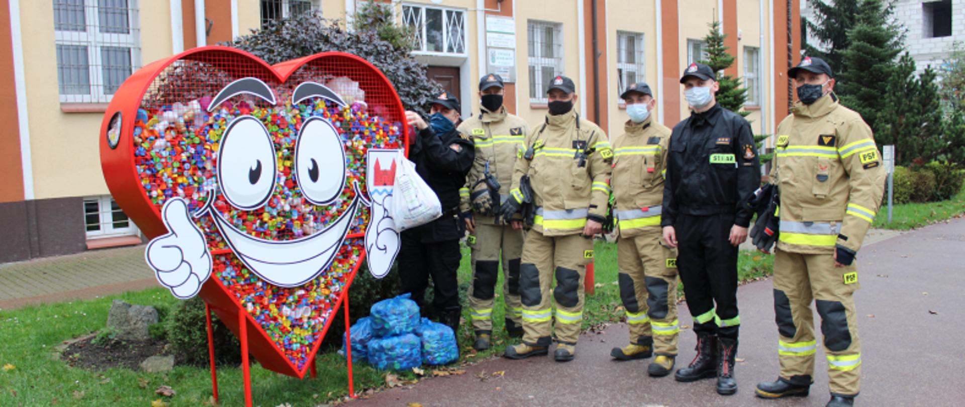 Zdjęcie sześciu funkcjonariuszy JRG Komendy Powiatowej PSP w Nidzicy przy pojemniku w kształcie serca z zebranymi zakrętami do butelek wewnątrz. Strażacy stoją na chodniku. Obok nich stoją worki niebieski z nakrętkami do butelek. Czterech strażaków w ubraniu specjalnym w kolorze pisakowym w czapkach z daszkiem i w maseczkach ochronnych w kolorze czarnym i niebieskim. Dwóch strażaków w ubraniach dowódczo-sztabowych w kolorze czarnym w czapkach z daszkiem i w maskach. Jedne z nich zawiesza worek foliowy z zakrętkami na pojemniku do zbierania zakrętek. W tle budynek w kolorach pomarańczowym i brązowym i białym. Z czerwonym dachem. Na ścianie widoczne godło z orłem oraz tablica informacyjna. 