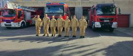 Na zdjęciu widać strażaków ubranych w mundury koszarowe, stojących w szeregu przed trzema czerwonymi wozami strażackimi. Jeden z nich, stojący jako pierwszy z lewej strony, oddaje salut. Strażacy stoją na placu przed budynkiem straży pożarnej — w tle znajduje się prosty, dwupiętrowy budynek z kilkoma oknami oraz trzema czerwonymi bramami garażowymi, z których wystają wozy strażackie.