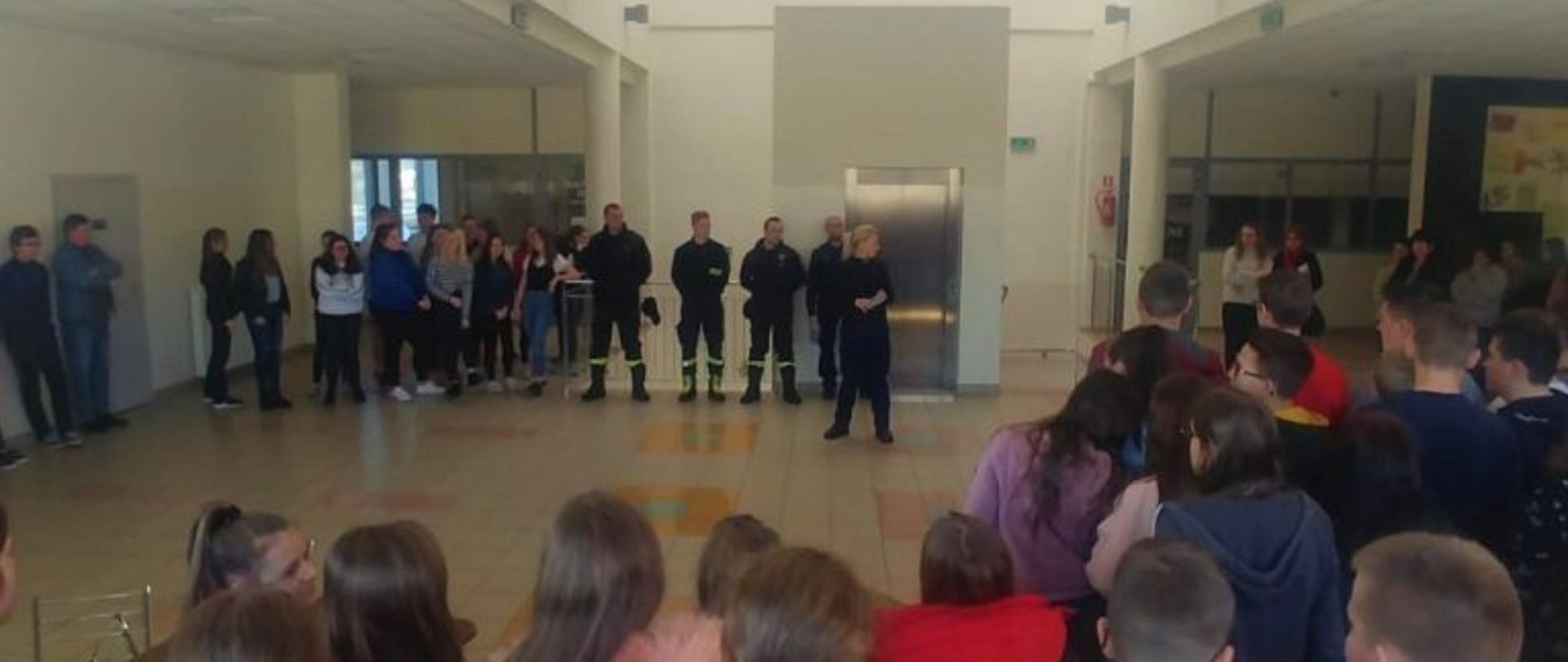 Zdjęcie przedstawia spotkanie funkcjonariuszy policji i straży pożarnej w umundurowaniu służbowym w pomieszczeniu. W spotkaniu uczestniczą dzieci. 