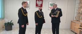 Trzech funkcjonariuszy ubranych w mundur galowy stojących w sali konferencyjnej. Jeden z nich trzyma w reku ozdobny dzwonek mosiężny na toporku z zamiarem przekazania go strażakowi. W tle na ścianie wisi godło Rzeczypospolitej. logo PSP oraz goło starostwa.