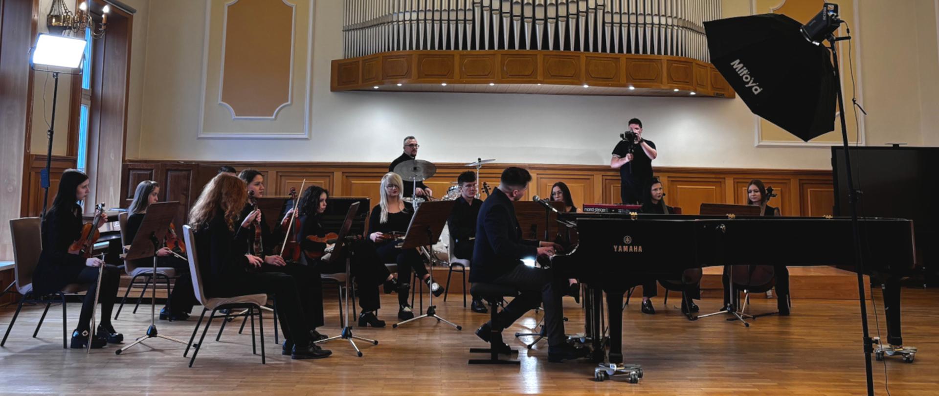 Na zdjęciu członkowie orkiestry szkolnej ZPSM oraz zasiadający przy fortepianie wokalista Krzysztof Iwaneczko. Scena ma miejsce na sali koncertowej ZPSM podczas kręcenia wideoklipu do piosenki Krzysztofa Iwaneczko. Na scenie znajduje się także kamerzysta oraz elementy profesjonalnego oświetlenia. Wszystkie obecne na scenie osoby ubrane są w galową odzież w kolorze czarnym. 