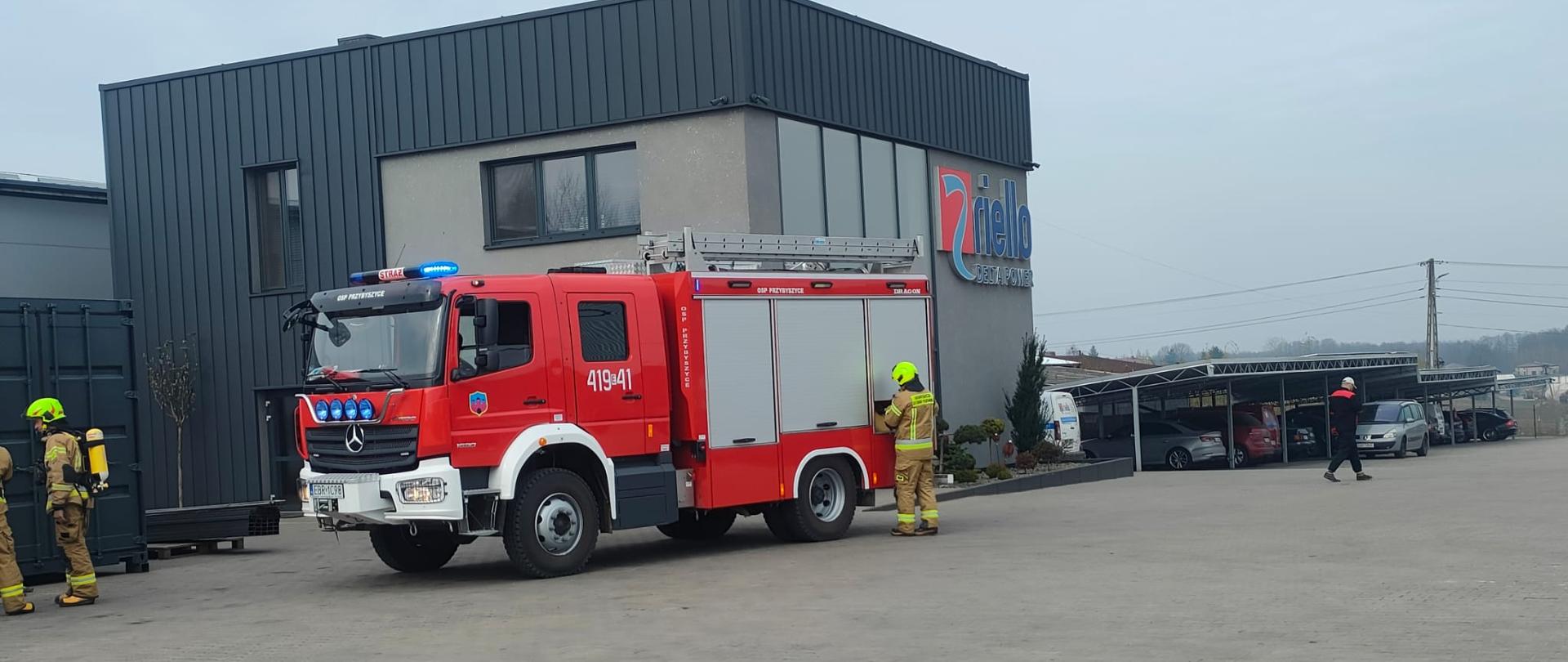 Ćwiczenia obiektowe w Jeżowie, samochód strażacki na placu firmy Riello zaparkowany przed budynkiem administracyjnym, dwóch strażaków przygotowuje się do wejścia do hali, w którym powstał pożar i znajduje się osoba poszkodowana, jeden ze strażaków wyjmuję sprzęt z skrytki wozu strażackiego.
