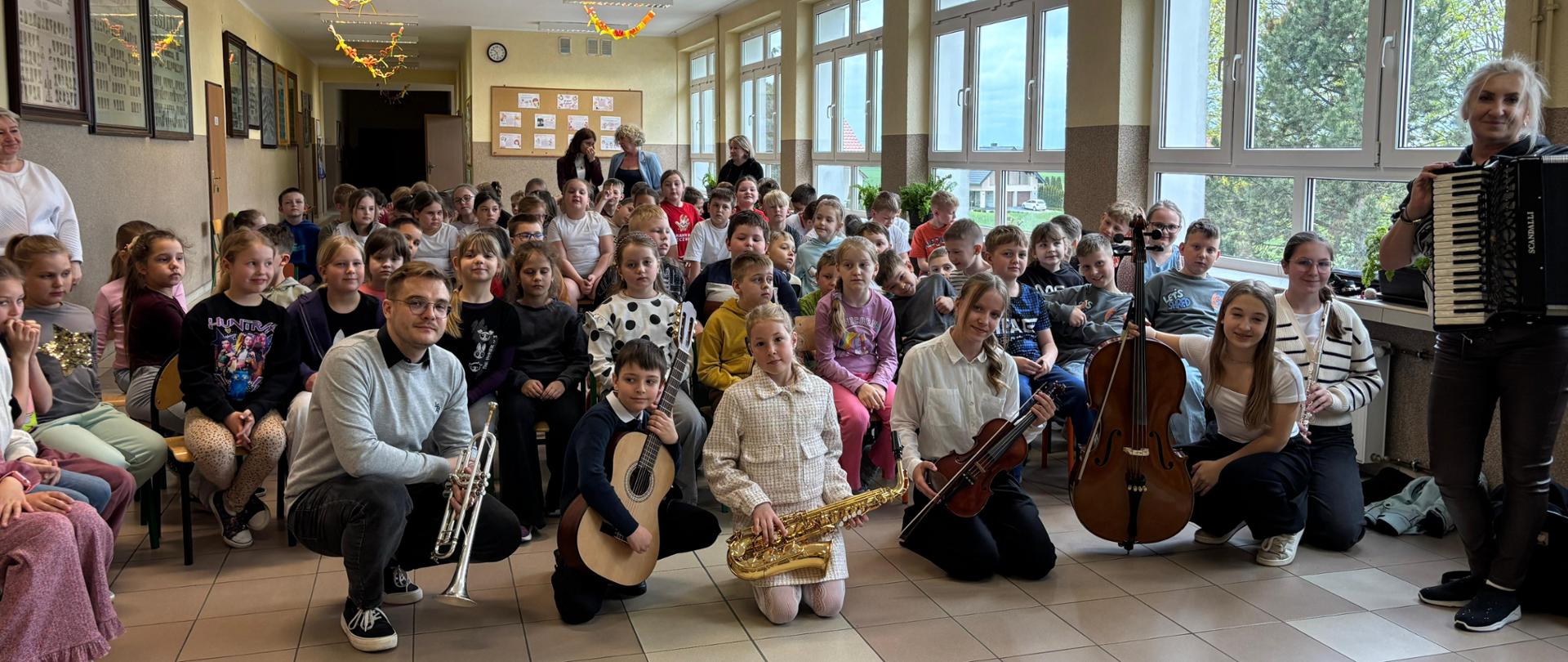 Młodzi muzycy z różnymi instrumentami pozują do pamiątkowego zdjęcia na środku szkolnego korytarza. W tle na krzesłach siedzi liczne grono uczniów obserwujących całe wydarzenie.