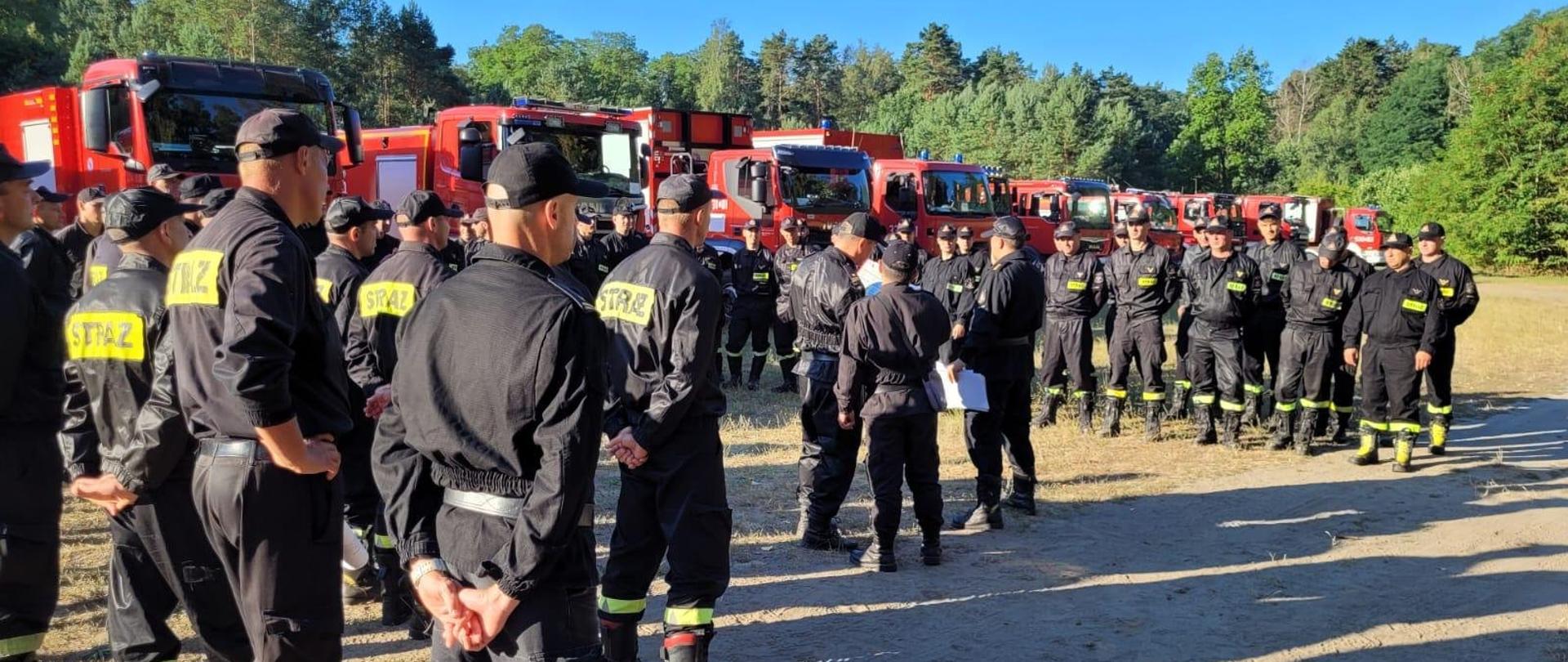 Na zdjęciu widoczni funkcjonariusze przygotowujący się do odprawy. W tle widoczne samochody pożarnicze, za nimi las.
