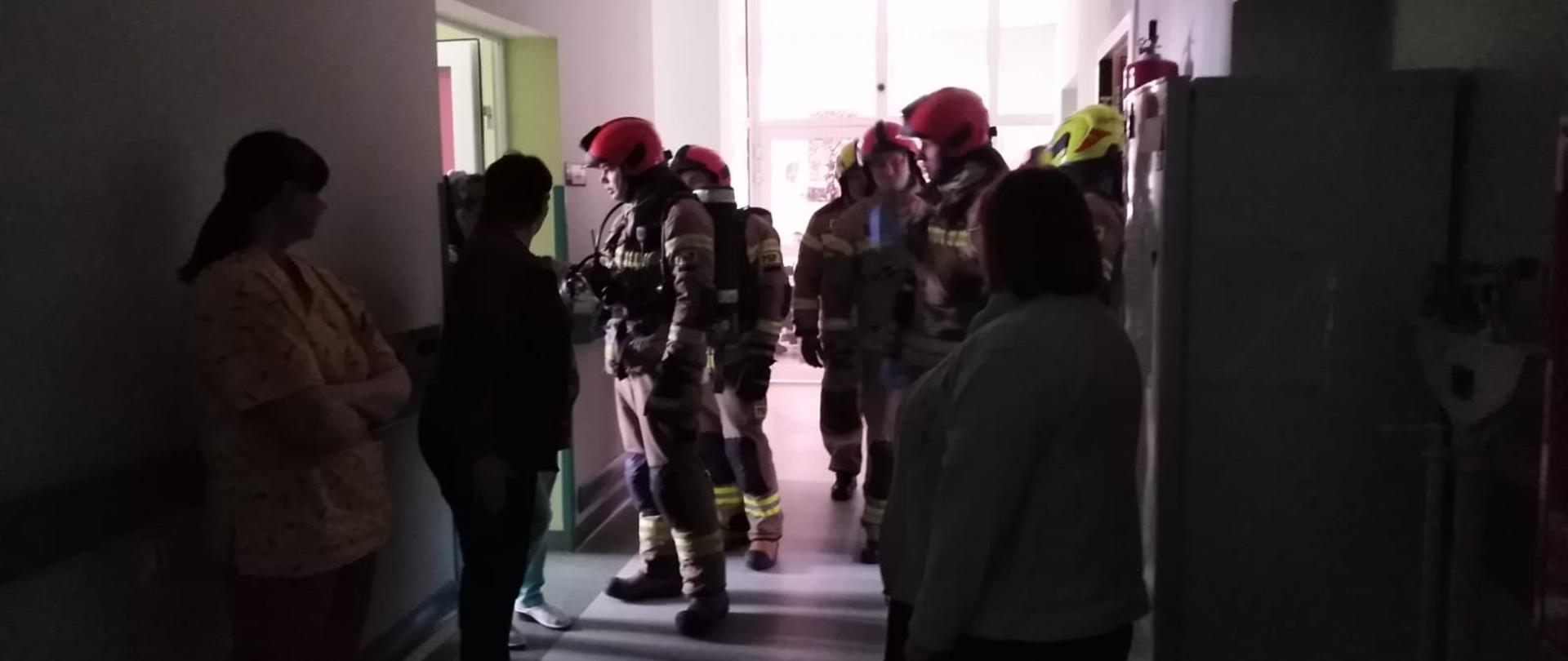 Zdjęcie przedstawia strażaków przygotowujących się do ewakuacji pacjentów. Widać korytarz budynku pięciu strażaków oraz trzy osoby personelu. 