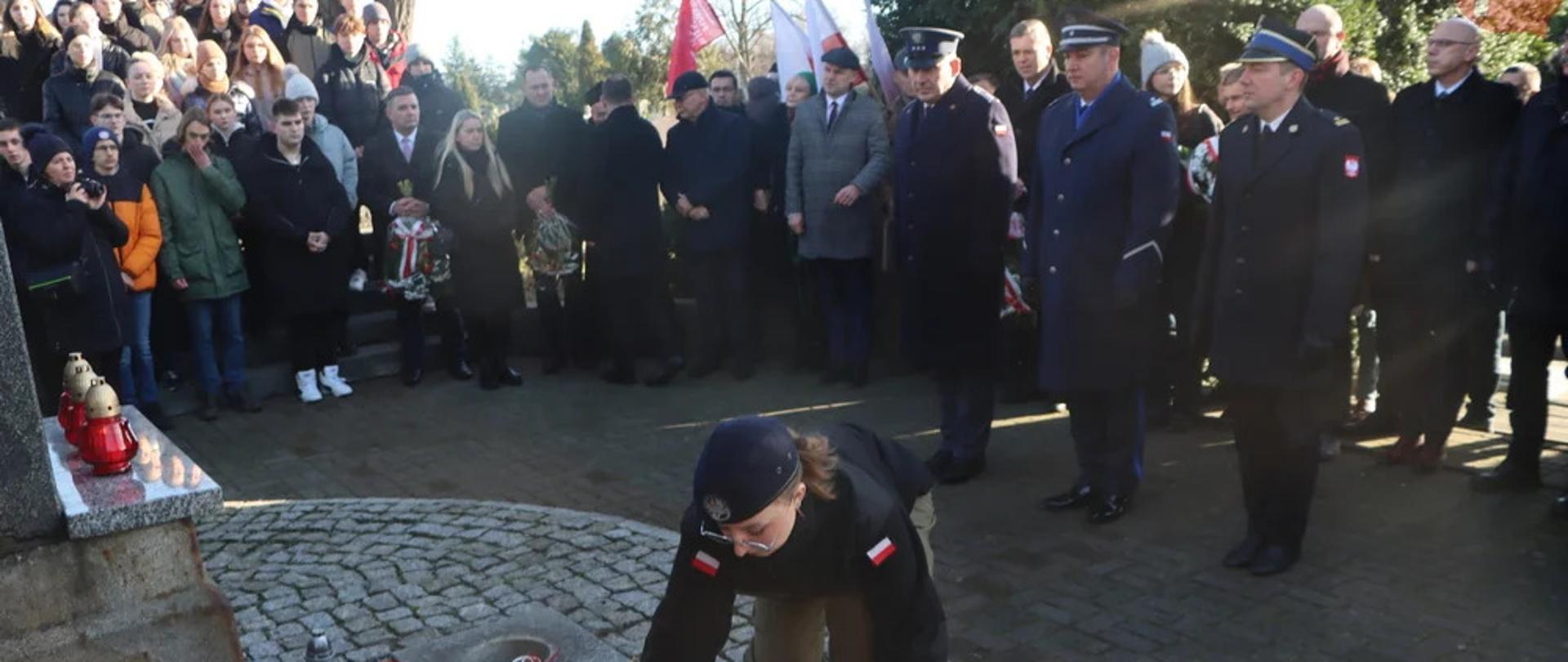 Cmentarz parafialny w Rawiczu, miejsce przed obeliskiem umapiętniającym Powstańców Wielkopolskich. Na przeciwko monumentu stoi 3 funkcjonariuszy: Państwowej Straży Pożarnej, Policji i Służby Więziennej - w umundurowaniu wyjściowym (zimowym). Uczennica klasy mundurowej składa wiązankę kwiatów przed tablica pamiątkową. Wokół inni uczestnicy uroczystości. W tle flagi i drzewa. 