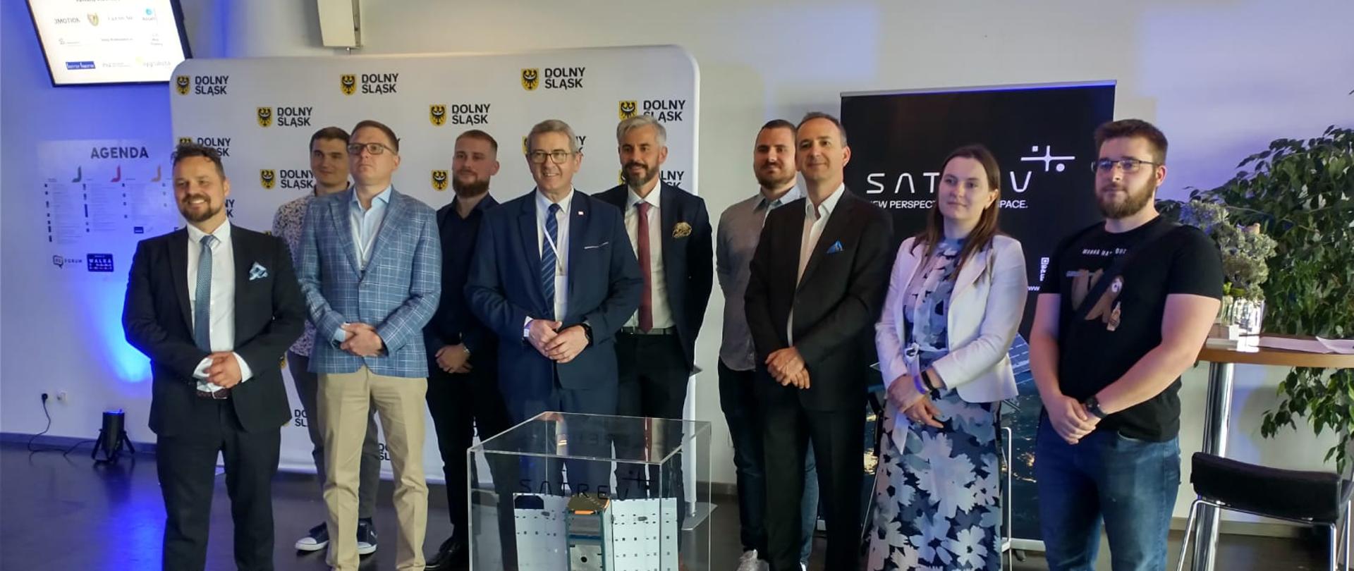 Grupa osób na tle baneru z herbem Dolnego Śląska. Po środku wiceminister Wojciech Murdzek. Przed nimi urządzenie w szklanej gablocie na stoliku. 