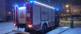 Strażacy PSP w ubraniu specjalnym stojący przy samochodzie ratowniczo-gaśniczym w porze nocnej przy obfitych opadach śniegu. Zdjęcie wykonane w przestrzeni miejskiej.