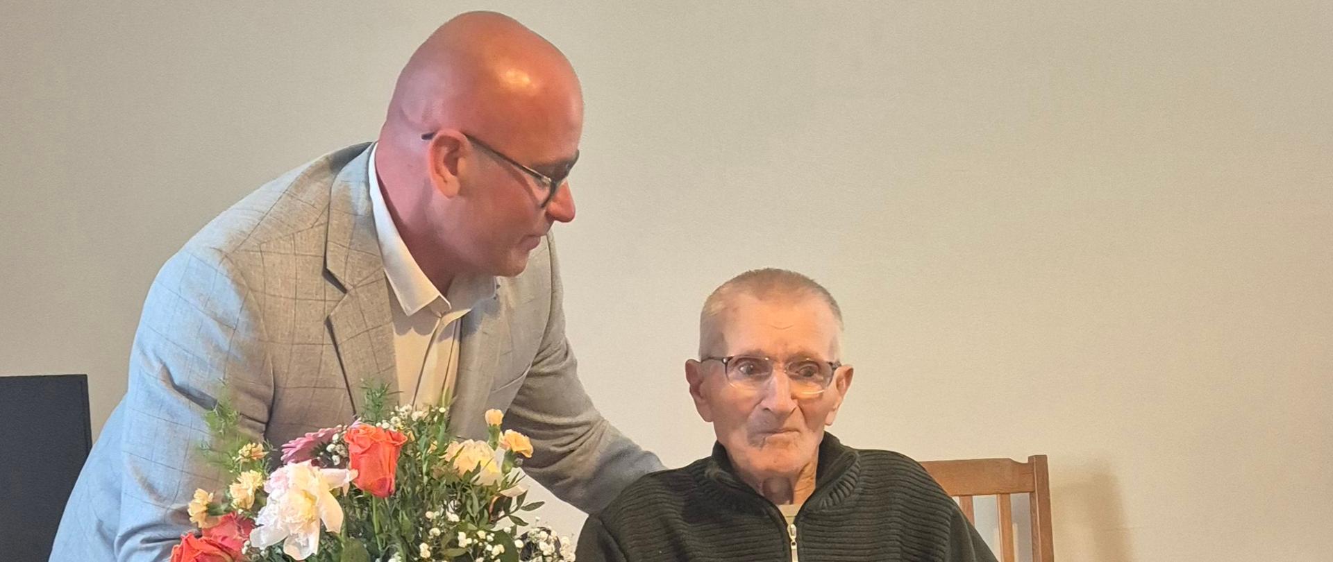 Starszy mężczyzna siedzi na wózku inwalidzkim i trzyma w rękach ozdobną zieloną teczkę ze złotym logo KRUS. Uśmiecha się do zdjęcia. Obok nachyla się nad nim młodszy mężczyzna z bukietem kolorowych kwiatów w ręce.