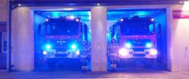 Dwóch strażaków stoi przy samochodach pożarniczych z włączonymi światłami samochodu stoją w garażu 