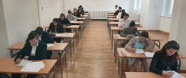 Zdjęcie przedstawia 18 uczniów klas najstarszych PSM w Głuchołazach, siedzą w oddzielnych ławkach ustawionych po bokach w białej sali kameralnej, przejście pośrodku. Uczniowie ubrani na galowo, ołówki w ręku, pochyleni nad kartkami z testem. 