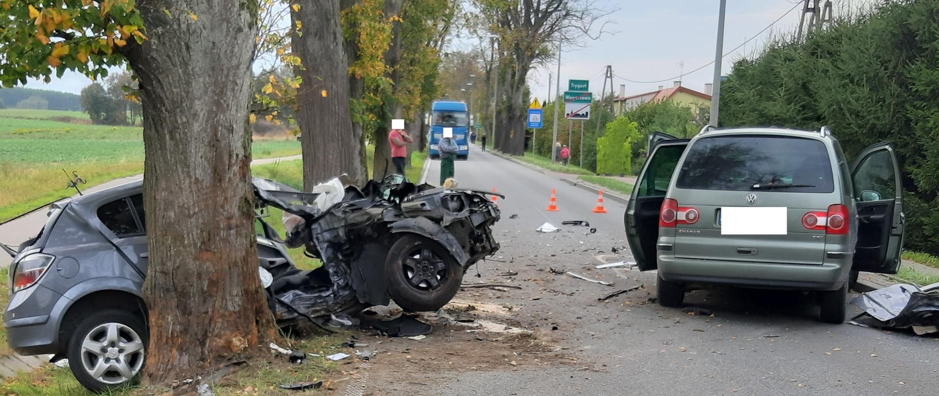 Zielony vw sharan z otwartymi tylnymi drzwiami oraz znajdujący się na skarpie szary opel astra z rozbitym całym przodem i bokiem. W tle niebieski samochód ciężarowy i 4 gapiów. 
