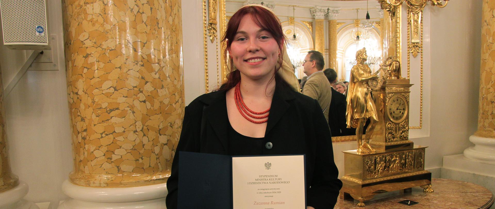 Na zdjęciu widzimy uczennicę Zuzannę Rumian trzymająca w rękach dyplom - Stypendium Ministra Kultury i Dziedzictwa Narodowego 2025. Fotografia została wykonana w Sali Balowej w Zamku Królewskim w Warszawie. Fot.: Marcin Klag