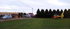 W prawej części zdjęcia widoczny śmigłowiec LPR stojący na trawie, w lewej części radiowóz, karetka pogotowia , samochody strażackie oraz strażacy. W tle zabudowania i drzewa iglaste - tuje.