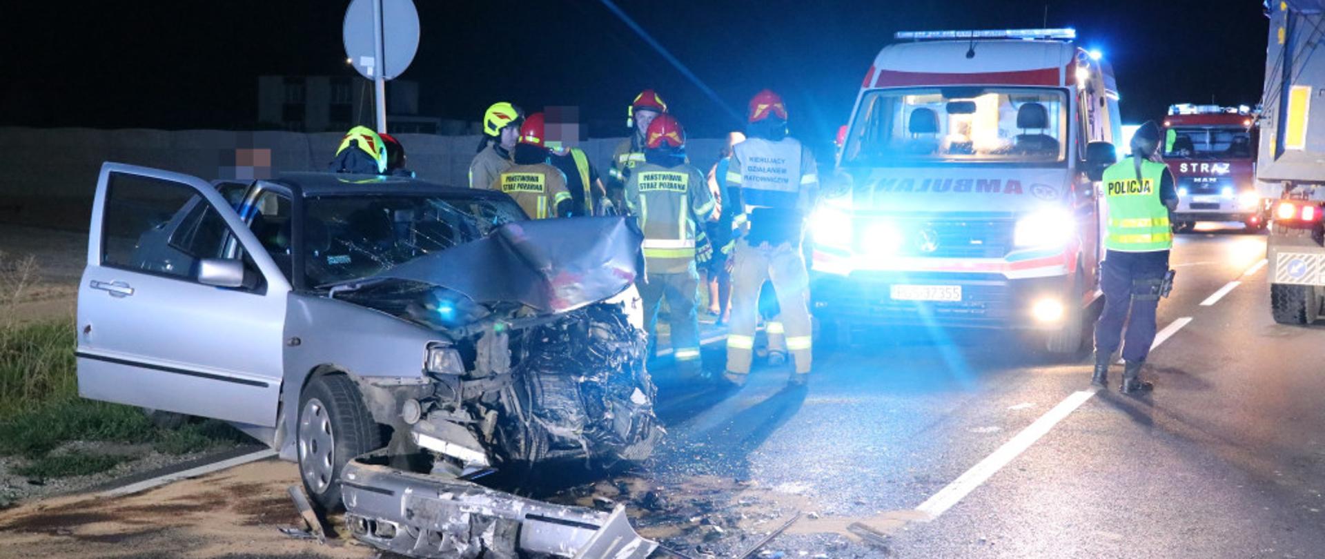 Na pierwszym planie widać uszkodzony samochód osobowy. W dalszym planie stoją strażacy oraz karetka pogotowia.