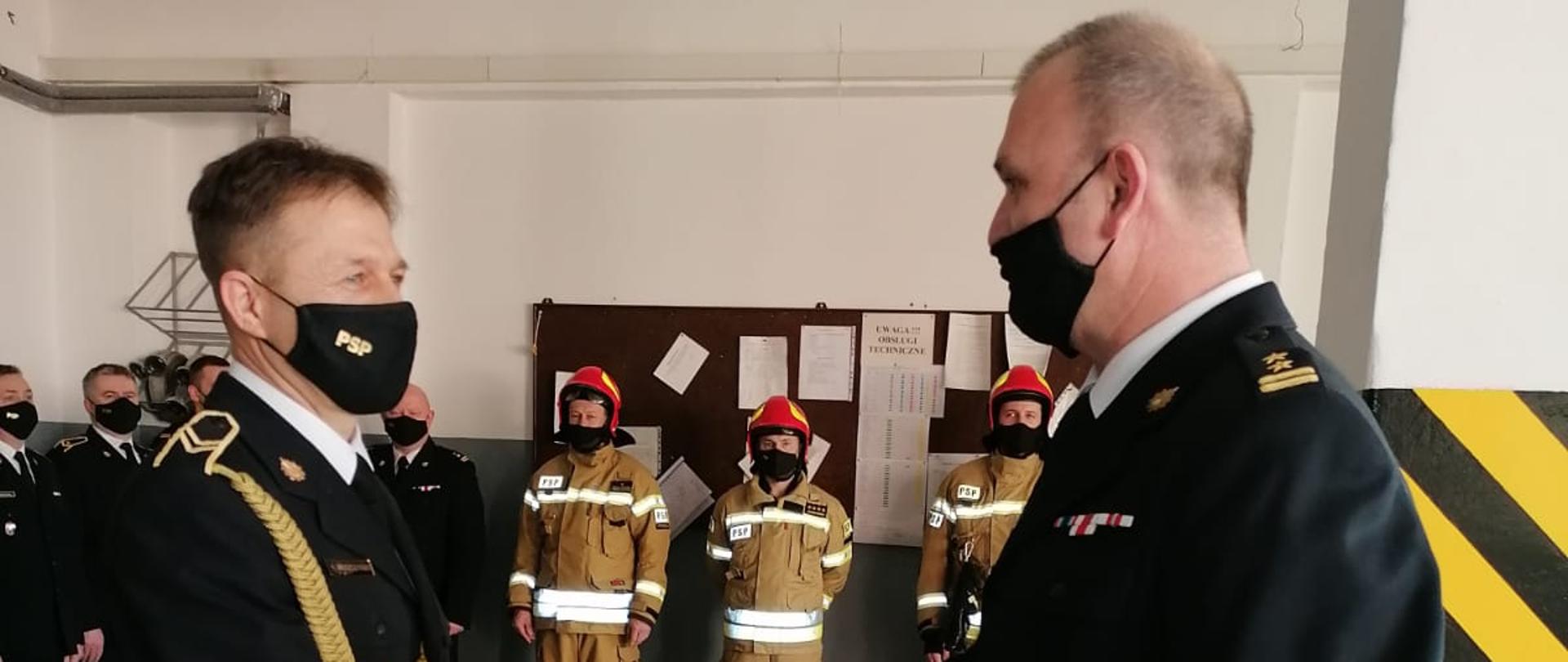 Zdjęcie przedstawia apel poświęcony funkcjonariuszowi , który przeszedł na zaopatrzenie emerytalne. Na zdjęciu widzimy funkcjonariusza żegnającego się ze służbą oraz Komendanta Powiatowego PSP w Ostrzeszowie. Zdjęcie wykonane w porze dziennej na garażu JRG.