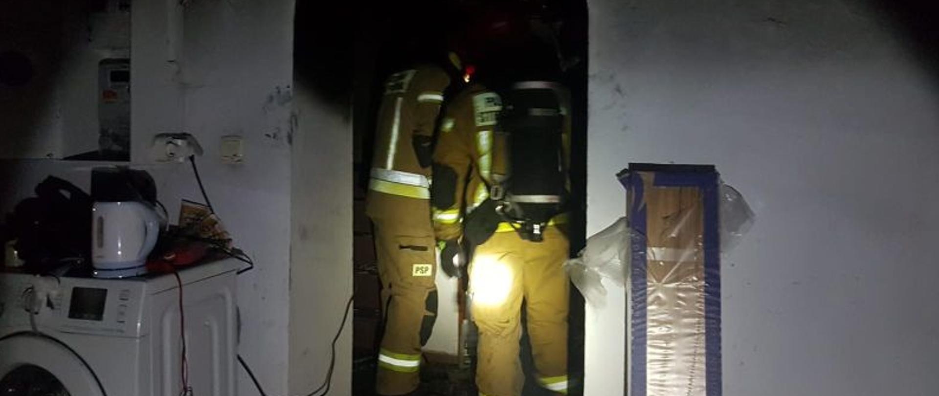 Centralny punkt zdjęcia stanowi dwóch strażaków widocznych od tyłu. Strażacy mają na sobie piaskowe ubrania i aparaty odo. Strażacy stoją w pomieszczeniu gdzie prowadzone były działania.