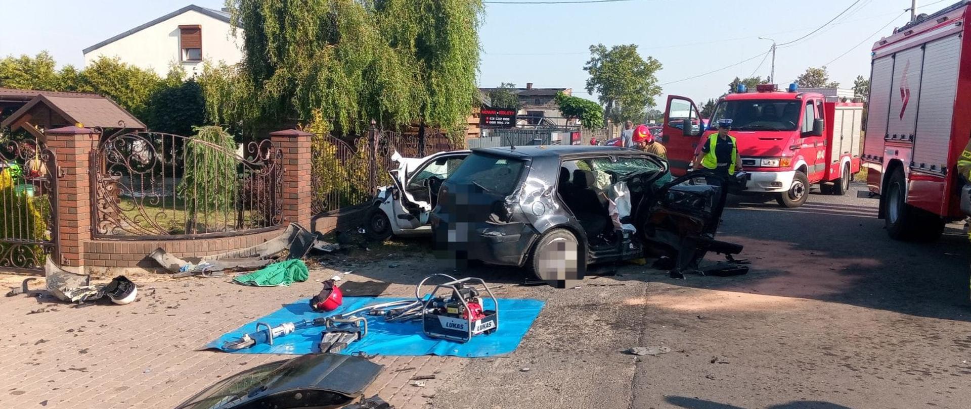 Na zdjęciu widać miejsce, w którym doszło do zderzenia dwóch samochodów osobowych. Biały i szary samochód są całkowicie uszkodzone. Obok leżą porozrzucane elementy karoserii i wyposażenia. Obok stoją dwa czerwone samochody strażackie. Widać strażaków oraz policjantkę. Na niebieskiej plandece na ziemi ułożone są strażackie narzędzia. Obok stoi czerwona torba medyczna. W tle widać budynki, płot, linie energetyczne oraz drzewa. Jest widno.