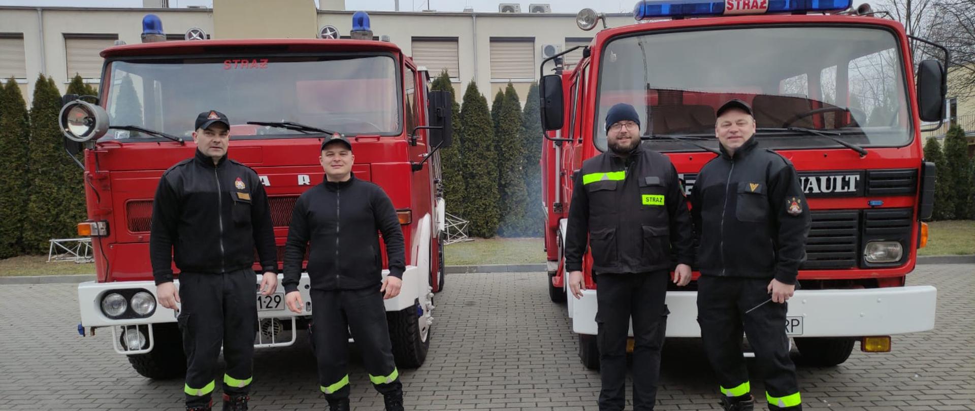 dwa samochody stoją na placu wyłożonym kostka przed nimi czterech strażaków w ubraniach koszarowych czarnych za nimi w tle zielone drzewa oraz budynek