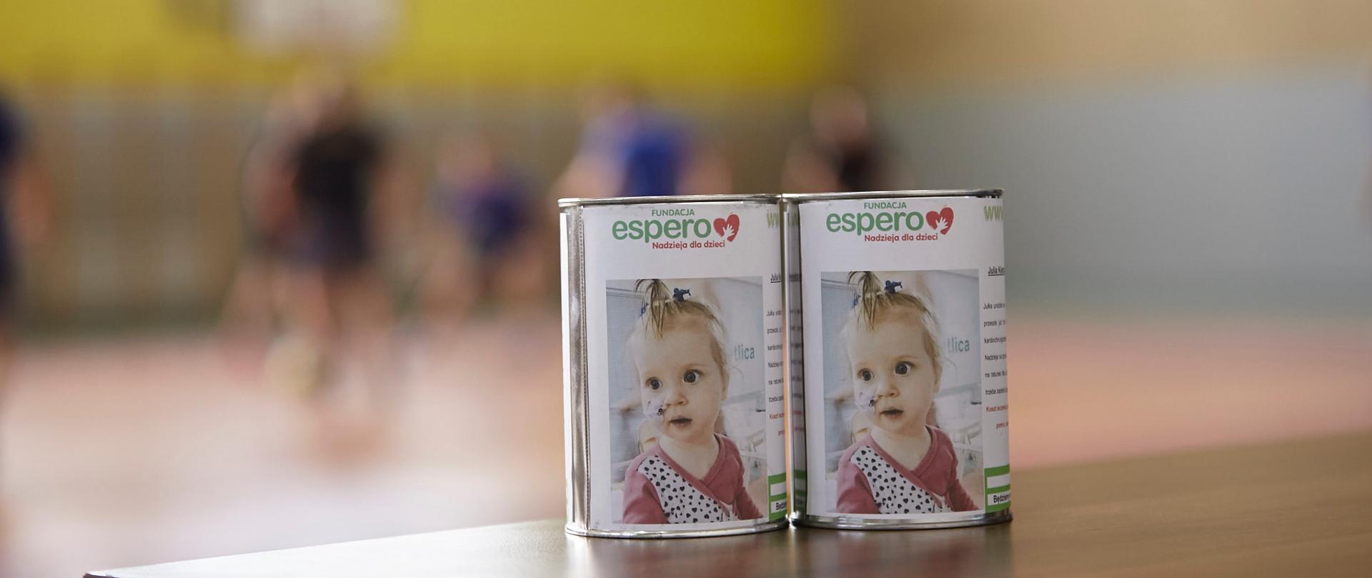Dwie puszki stojące na stole przeznaczone na zbiórkę pieniędzy. Na puszkach widoczne jest zdjęcie dziewczynki, na której rzecz zostaną one przekazane. 