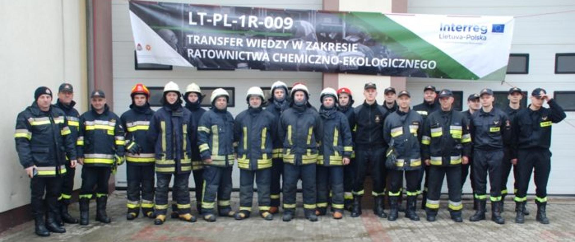 Strażacy u ubraniach specjalnych ustawieni w szeregu na tle wrót garażowych 