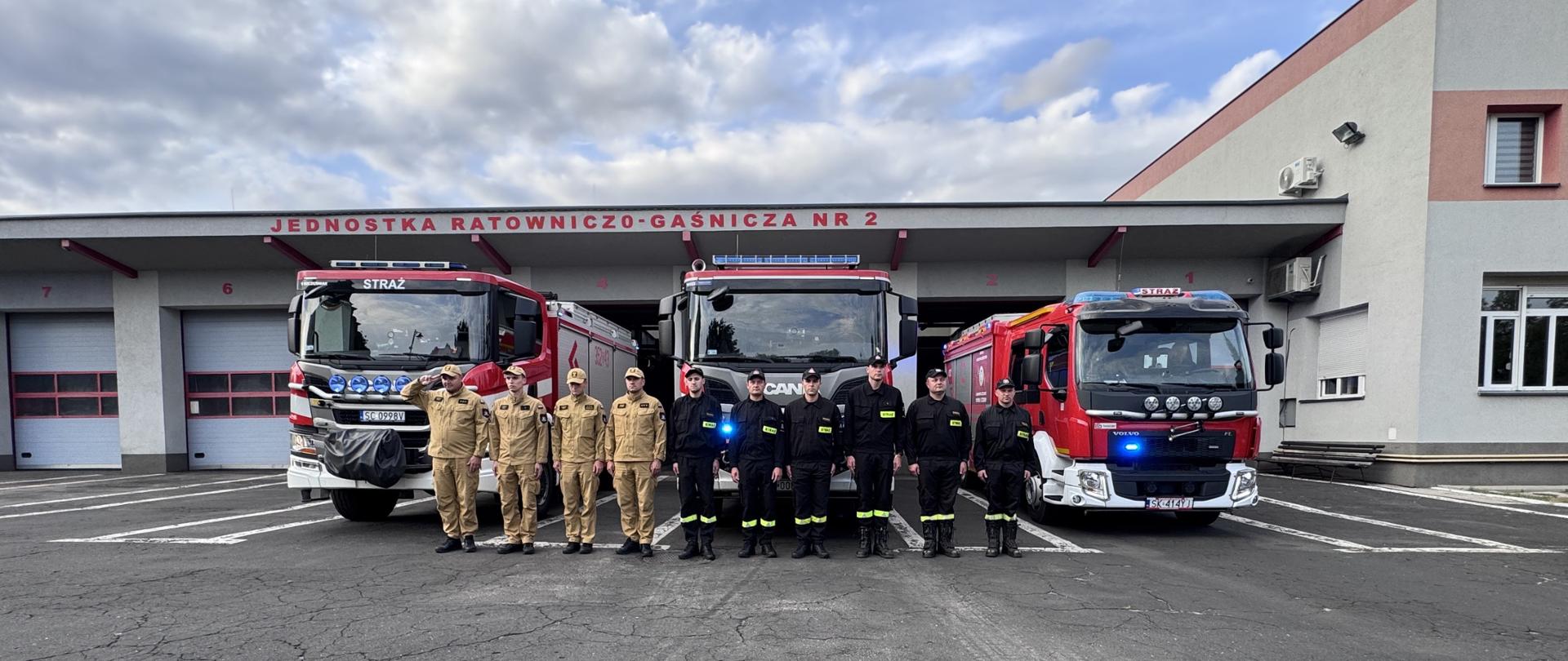 Na zdjęciu widzimy dziesięciu strażaków stojących przed budynkiem remizy i trzema wozami strażackimi. Budynek, w jasnym kolorze z czerwonymi akcentami, ma trzy bramy garażowe z napisem "JEDNOSTKA RATOWNICZO-GAŚNICZA NR 2" nad nimi. Od lewej strony, w bramach widoczne są wozy oznaczone numerami 6, 3 i 7.
Strażacy ustawieni są w rzędzie, zwróconymi w stronę obiektywu. Sześciu z nich, po lewej stronie, ubranych jest w jasne, piaskowe mundury, z kolei czterech, po prawej, nosi ciemne, granatowe lub czarne uniformy. Mają na sobie kurtki i spodnie. Na tle nieba, które jest częściowo zachmurzone, widać fragmenty drzew i dachu budynku po prawej stronie. Po prawej stronie zdjęcia znajduje się także duża, szara ściana z oknami, stanowiąca część remizy.