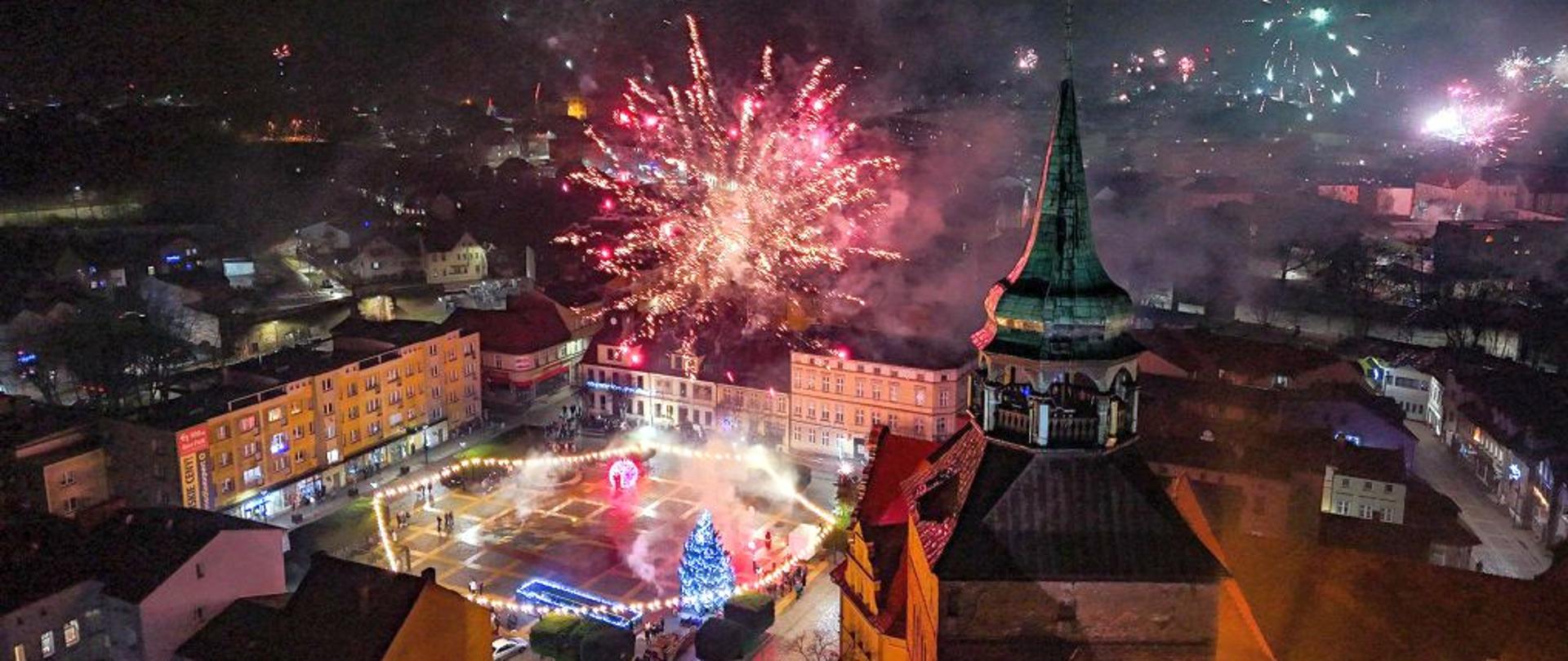 Widok na miasto w noc sylwestrową. Zdjęcie rozświetlają fajerwerki.