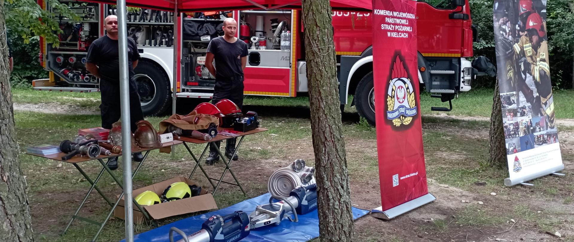 Zdjęcie przedstawia miejsce prezentacji sprzętu straży pożarnej. Na pierwszym planie stoisko z prezentowanym sprzętem pożarniczym a za nim stoi samochód strażacki.