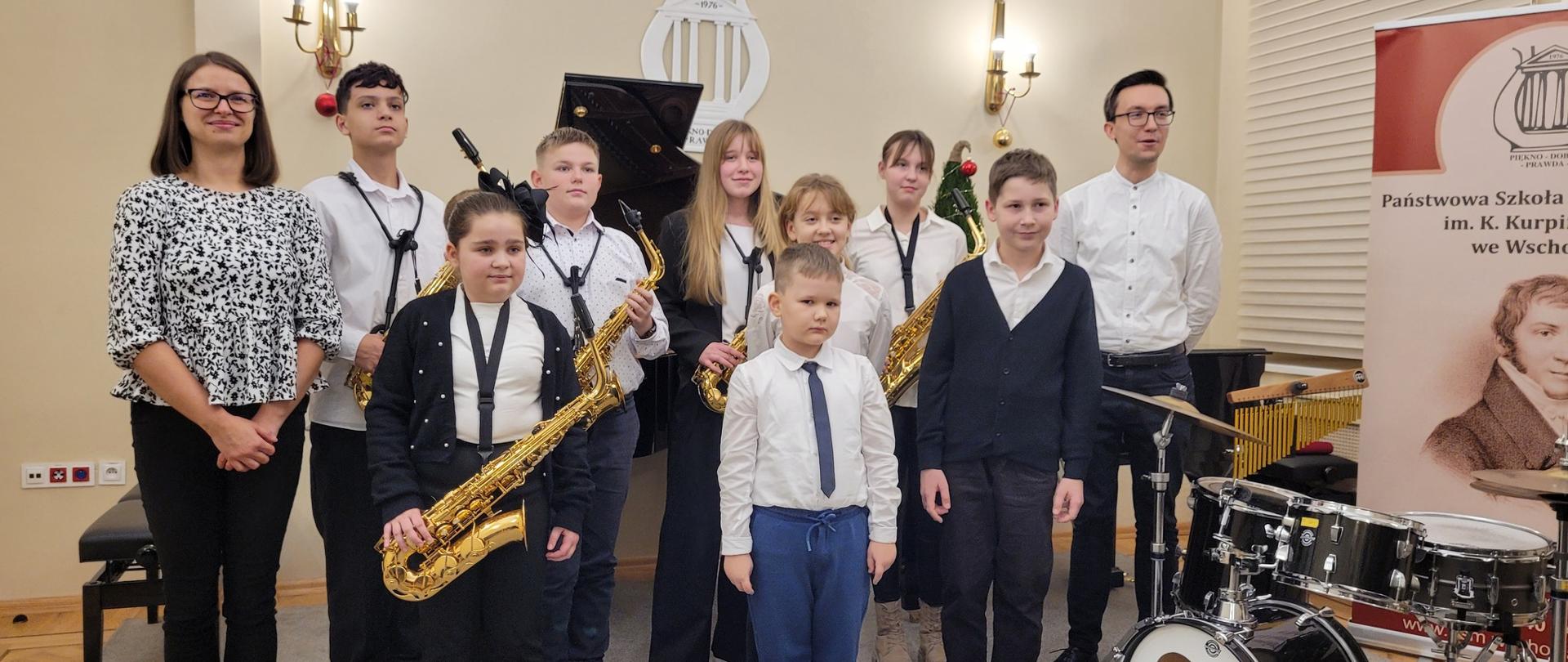 Zdjęcie uczniów występujących na koncercie oraz ich nauczycieli w auli szkoły.