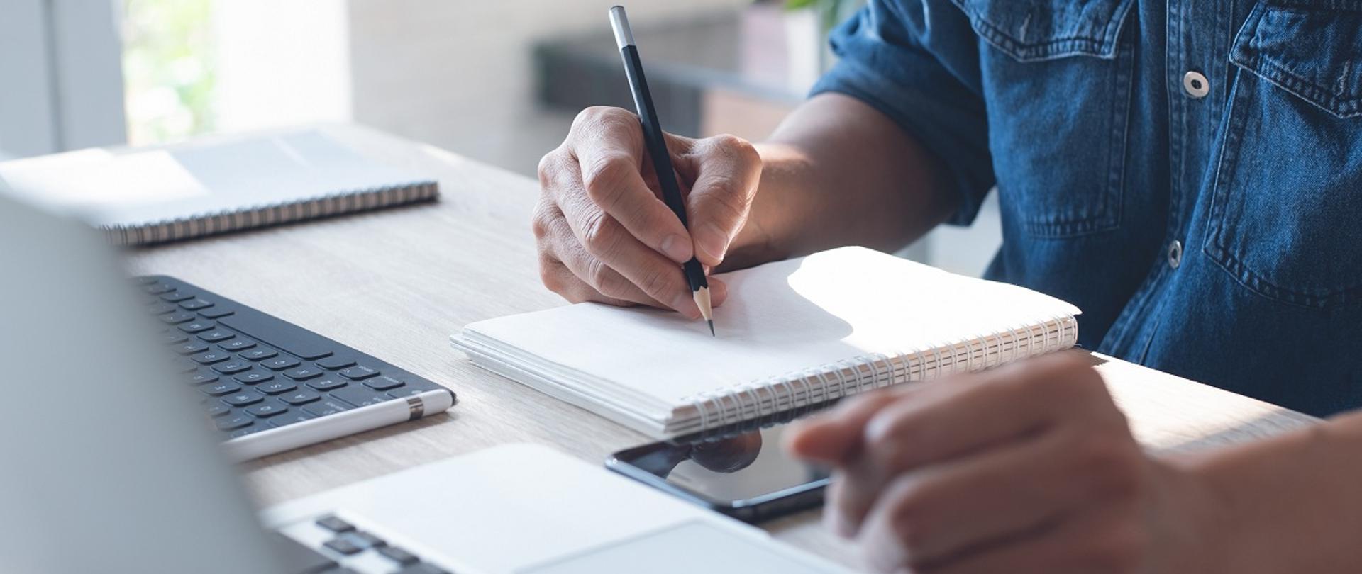 Dłonie na biurku. W jednej dłoni ołówek. Na biurku leży notes, telefon komórkowy i dwa laptopy.