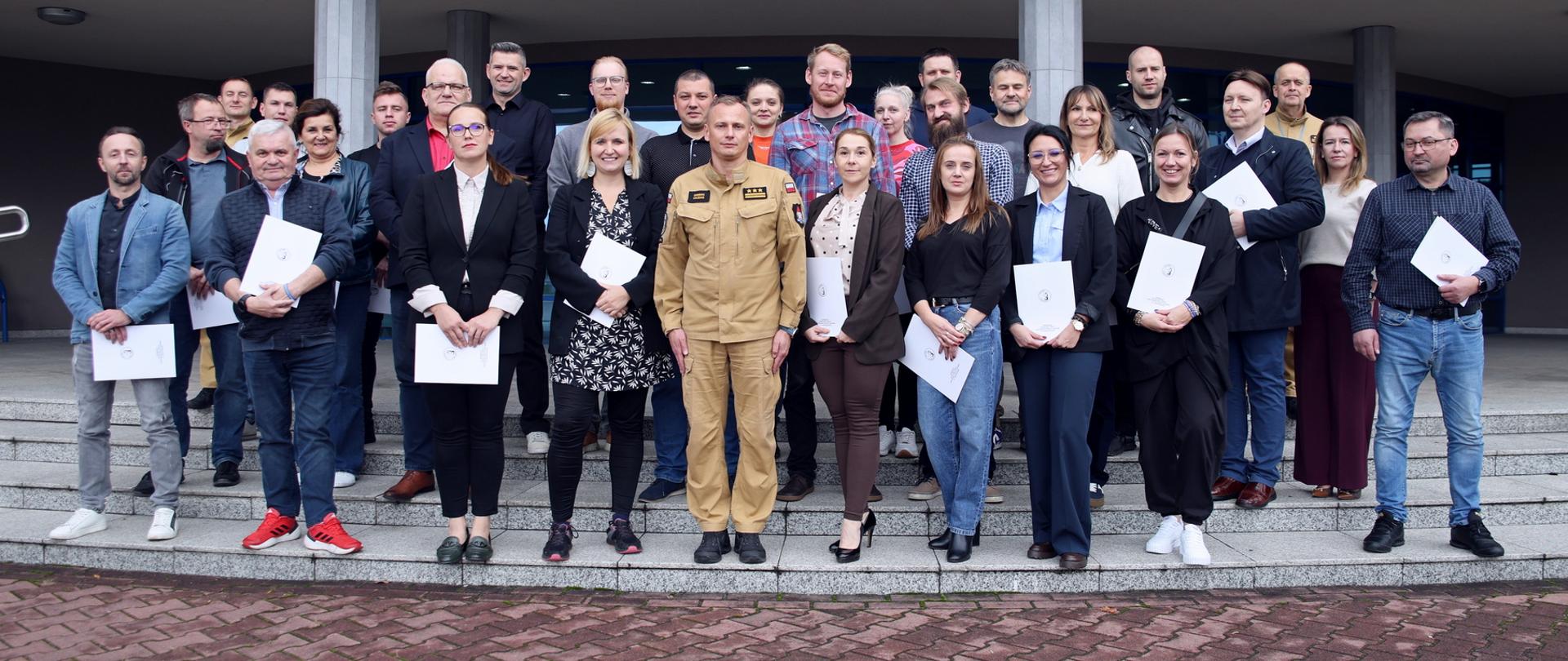 Pamiątkowe zdjęcie grupowe uczestników szkolenia inspektorów ochrony przeciwpożarowej (26 osób) w towarzystwie Zastępcy Komendanta Centralnej Szkoły PSP, kobiety oraz dwóch funkcjonariuszy CS PSP