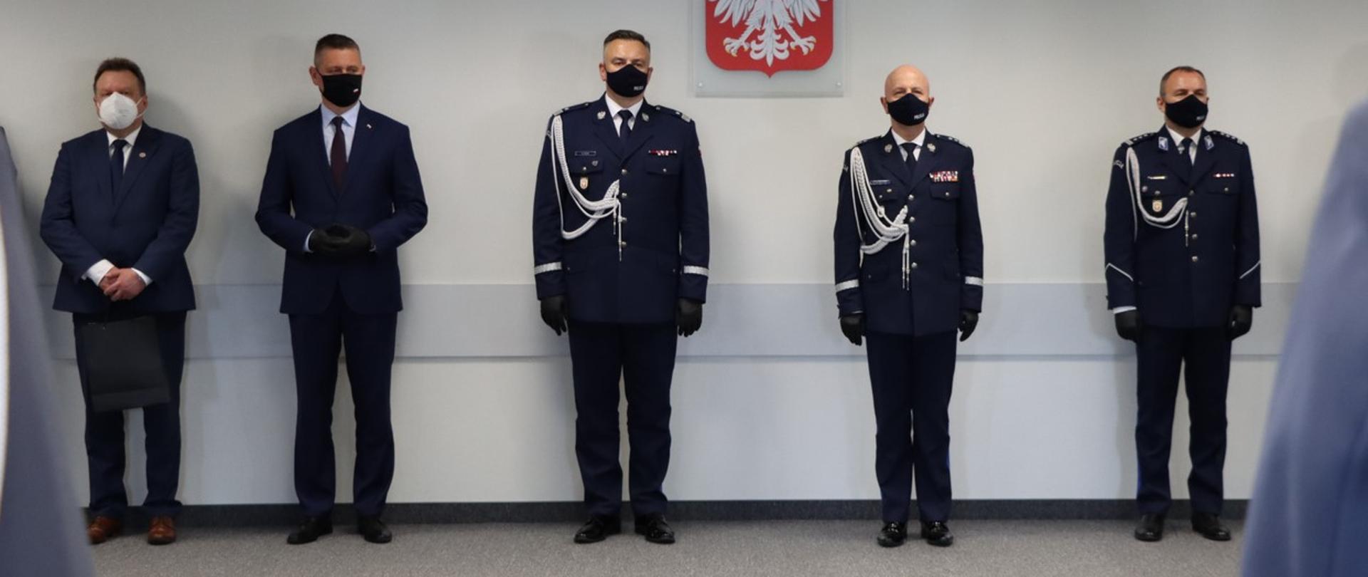 Na zdjęciu znajdują się mężczyźni ubrani w ciemne garnitury oraz policjanci w mundurach. Wszyscy są ustawieni w rzędzie.