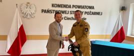 Zdjęcie przedstawia uroczysty moment przekazania sprzętu ratowniczego w siedzibie Komendy Powiatowej Państwowej Straży Pożarnej w Ciechanowie. Po lewej stronie stoi Prezydent Ciechanowa w beżowym garniturze, który ściska dłoń Komendantowi Powiatowemu PSP w Ciechanowie. Strażak trzyma w drugiej ręce hydrauliczne narzędzie ratownicze. W tle widoczne są dwie flagi Polski, godło państwowe oraz tablica z nazwą jednostki.